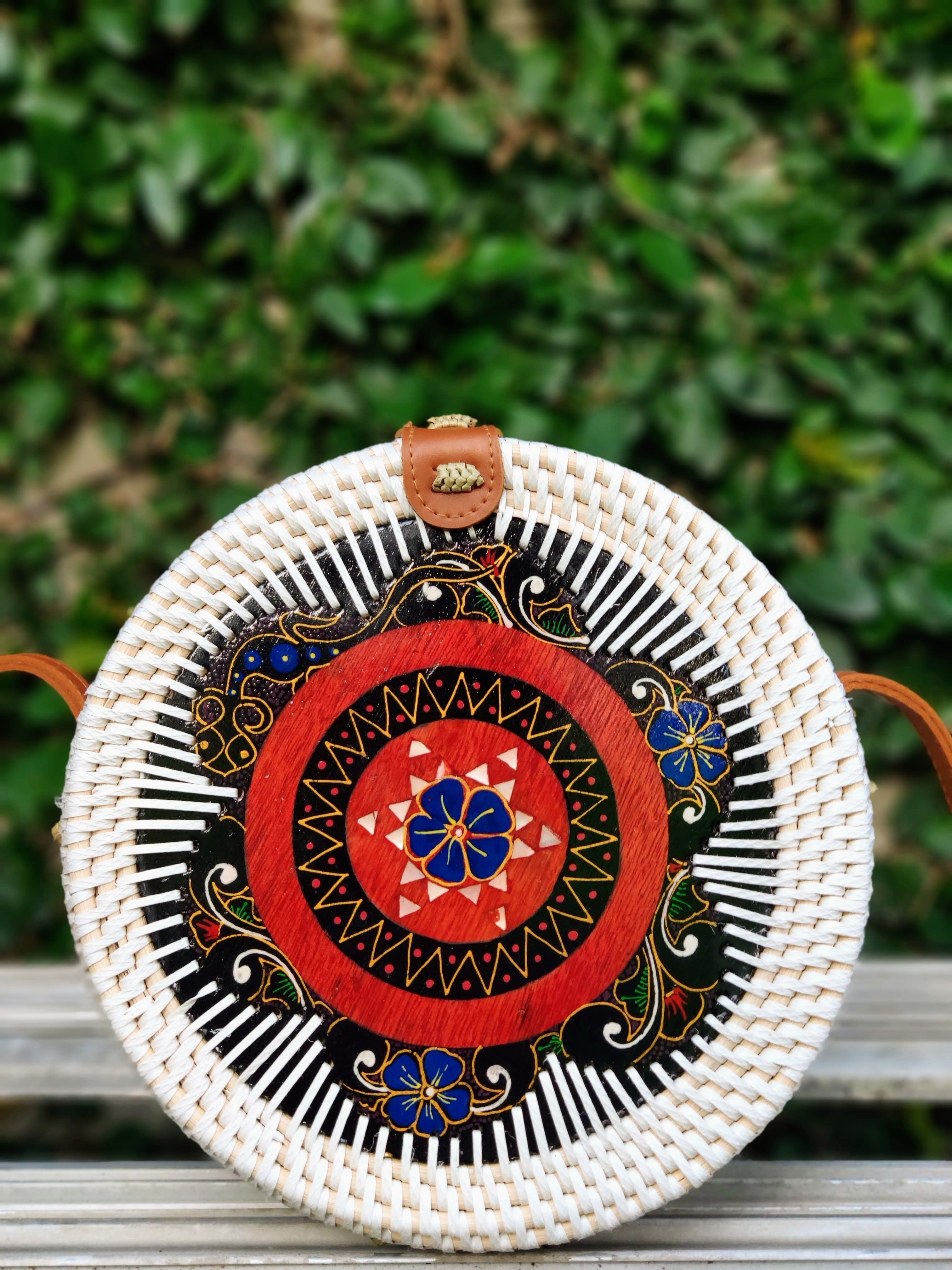 Round woven bag with decorative patterns on a wooden surface with green foliage in the background